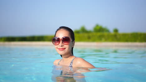 Sexy-Mujer-Asiática-Con-Grandes-Gafas-De-Sol-De-Pie-En-La-Piscina,-Sonriendo-A-La-Cámara