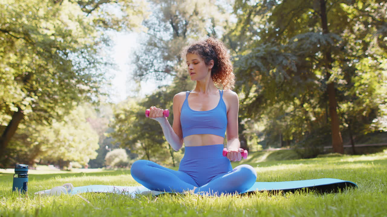Sportive woman exercising weightlifting with dumbbells sitting on yoga mat in park on sunny day