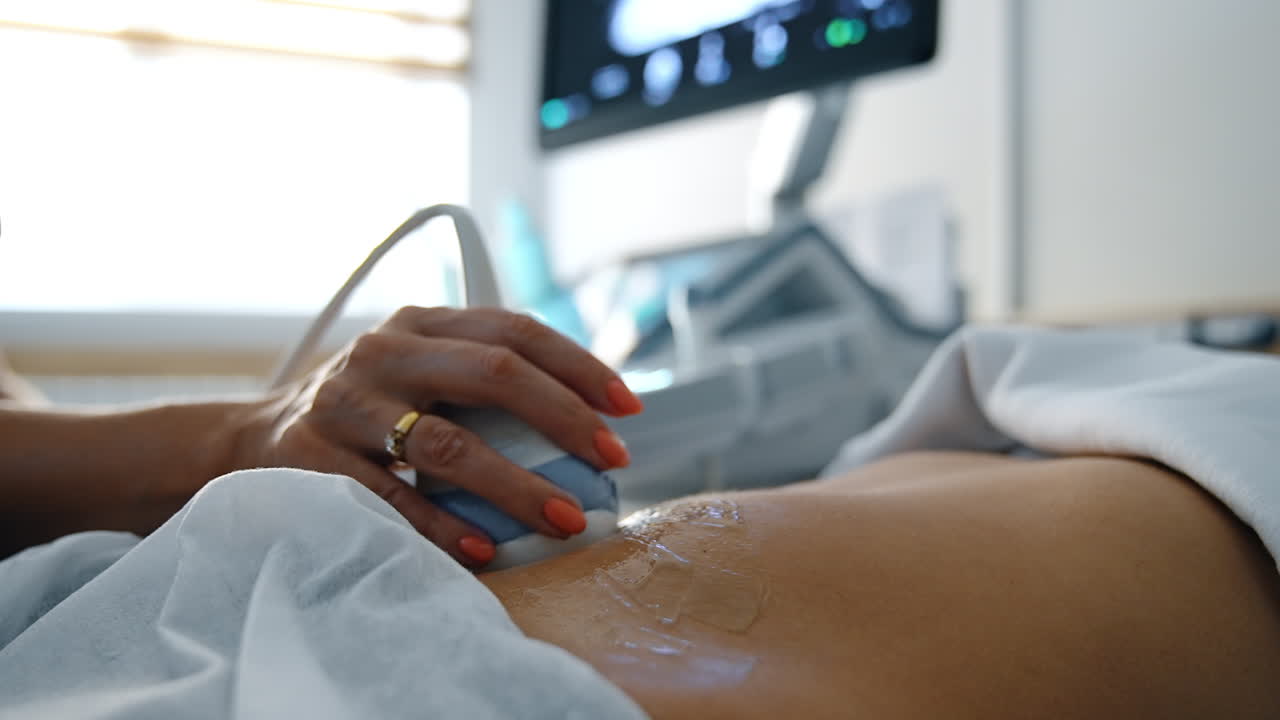 Doctor moving the ultrasound sensor by the gelled belly of pregnant patient. Close up. Monitor of machine at backdrop in blur.
