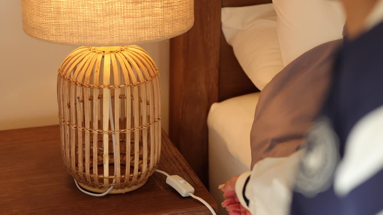 A person turns off a woven lamp on a bedside table, creating a warm, relaxed atmosphere in a cozy bedroom