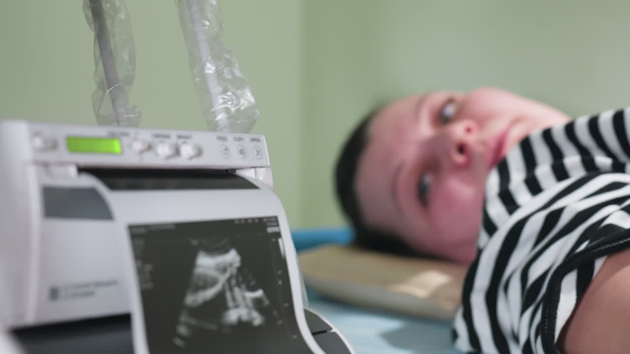 patient lying faced up on clinical bed turns to watch printer as lab result prints out on machine showing ultrasound image while she smiles warmly in response to progress of examination procedure