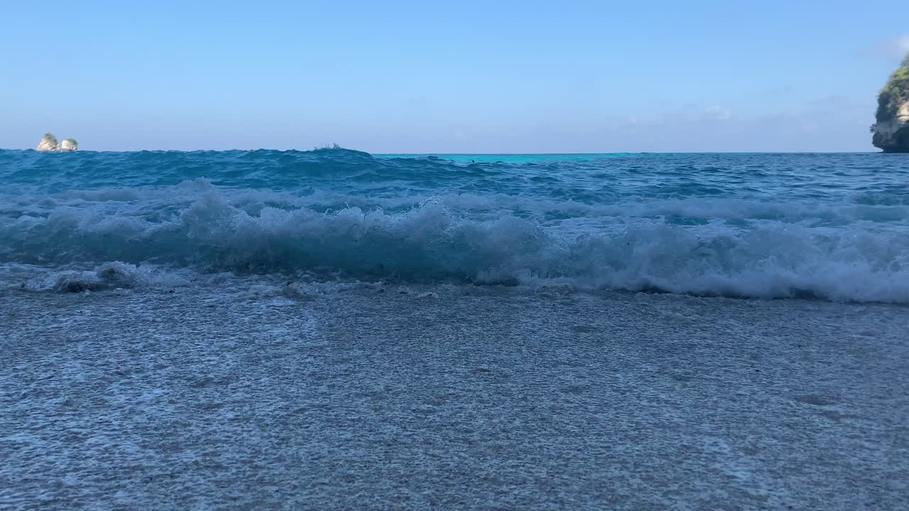 Close Up shot of turquoise blue waves washing ashore tumbling rocks with white sand beach in Diamond Beach, Nusa Penida, Bali, Indonesia.