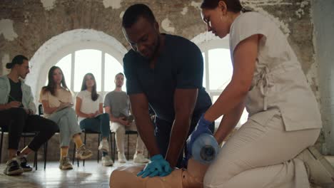 close-up shooting: a female doctor in a white uniform conducts a medical first aid course for the public, who listens by showing artificial respiration on the mannequin using the ambu resuscitation bag and explains his actions, a black male nurse in a blue uniform performs artificial respiration by