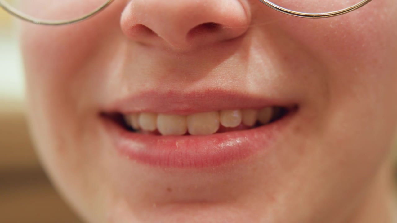 Primo piano di donna caucasica che sorride con una progressione delicata, insegnante che offre un sorriso gentile con occhiali visibili, labbra che passano da neutre a un sorriso ampio, illuminazione interna calda, texture naturale della pelle e denti rivelati