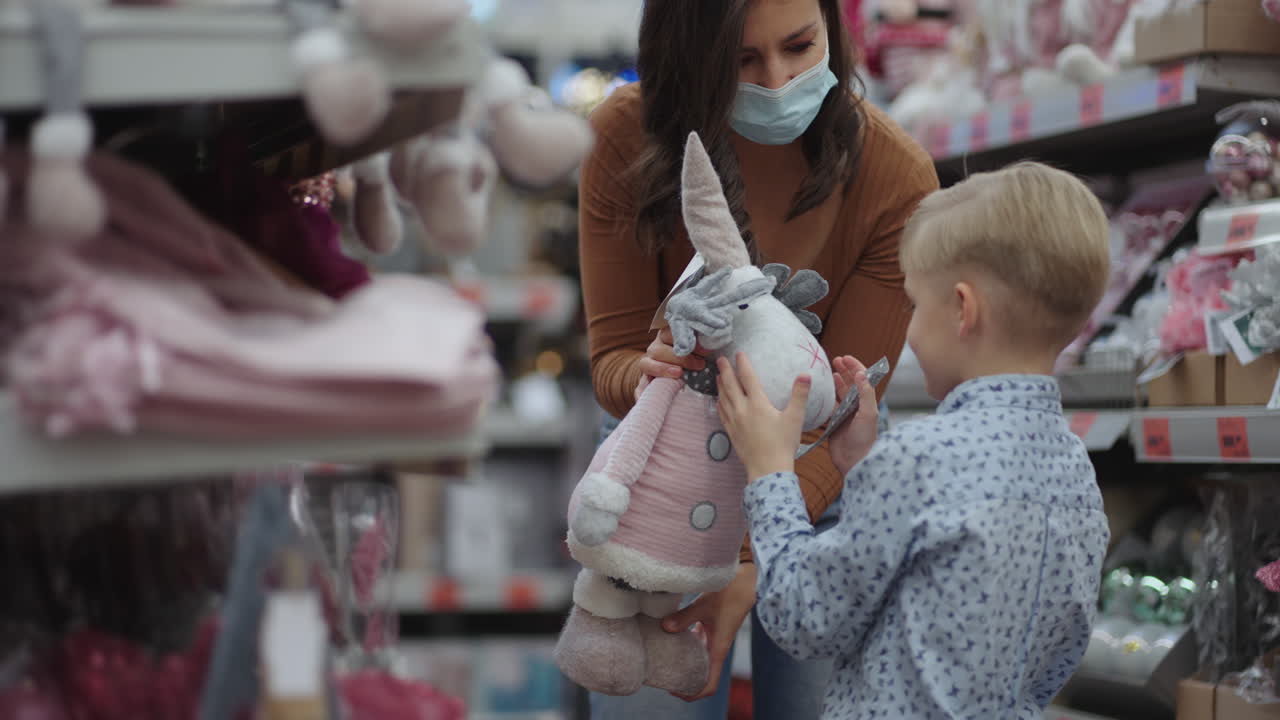 mujer joven con máscara protectora con su lindo hijo eligiendo papel de regalo de navidad en una tienda. compras navideñas y de regalos