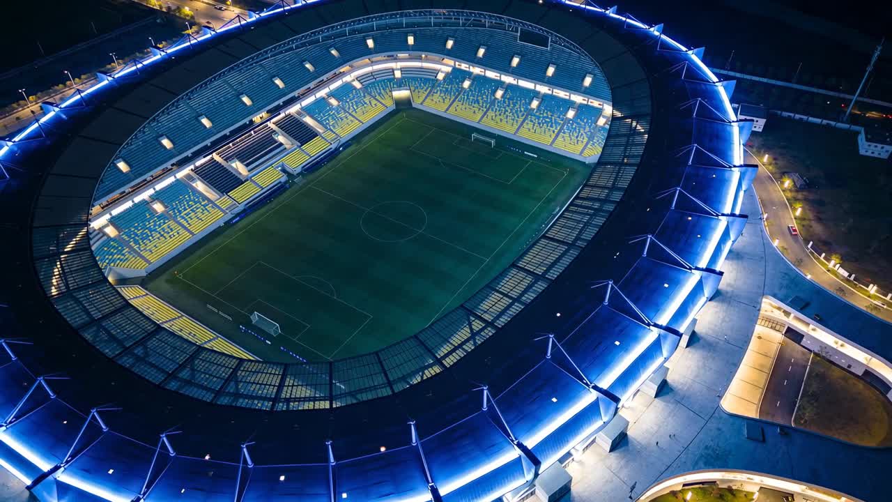 Drone che plana e ruota mostrando uno stadio ovale illuminato di notte, campo e porte vuoti, anello LED blu