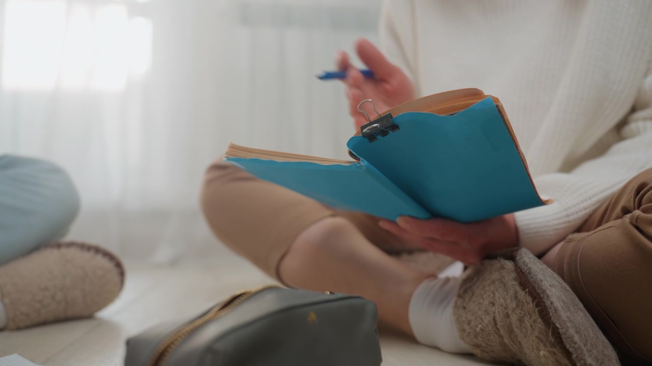 Mani caucasiche che scrivono su un quaderno, compagni di studio seduti a gambe incrociate sul tappeto che condividono un'agenda blu e una matita, astuccio grigio e pagine sparse, luce diurna soffusa attraverso le tende, mani che si scambiano appunti