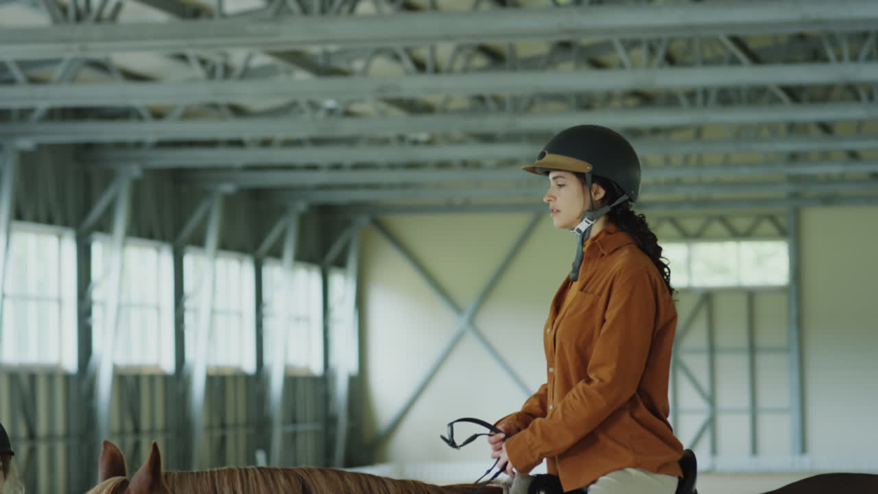 A woman riding a horse in an indoor equestrian arena with an instructor