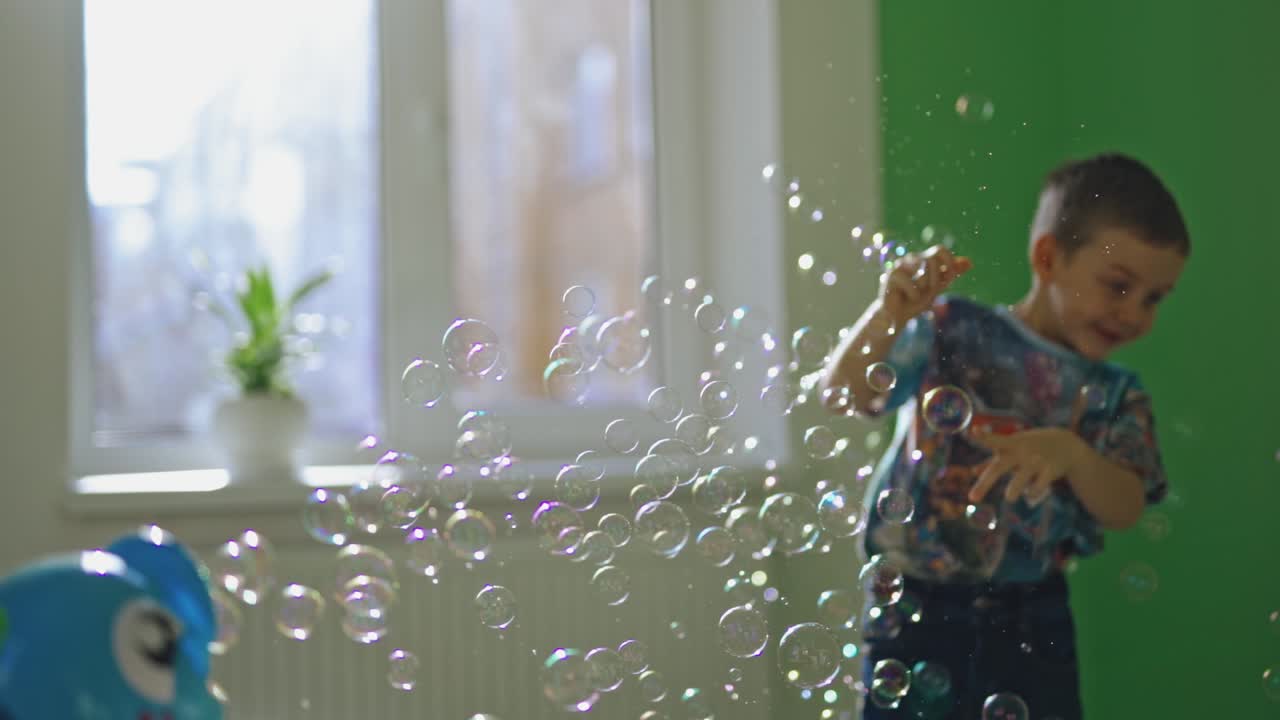 Joyful boy plays with soap bubbles in his room. Little child is popping colorful bubbles with his fingers and smiling at home.