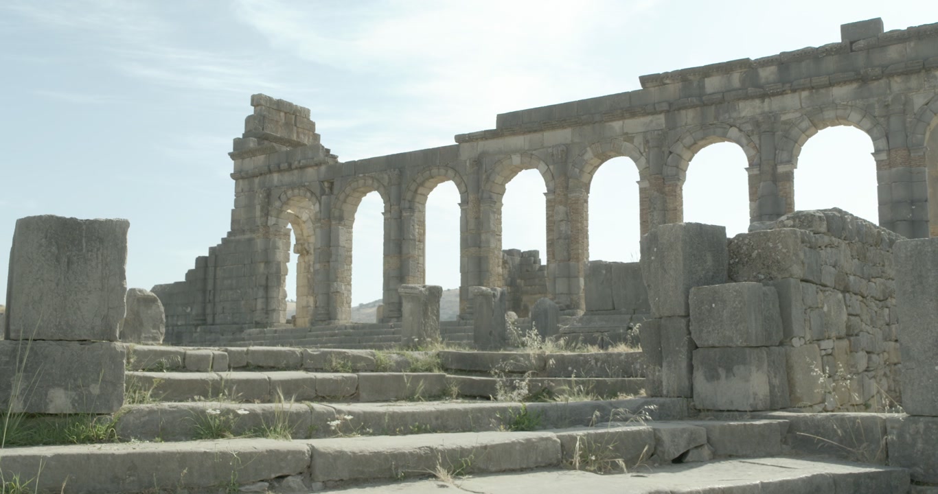 Vista statica a livello del suolo di gradini in pietra che conducono alla Basilica porticata di Volubilis, evidenziando l'architettura civile romana nel sito archeologico patrimonio dell'UNESCO del Marocco