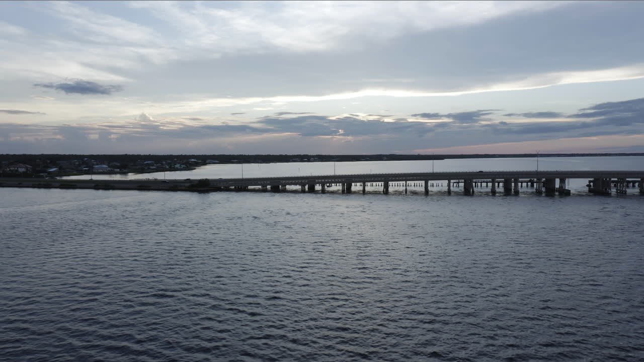 Gentle waves ripple beneath a long bridge spanning a tranquil bay, as the fading daylight reflects silver and blue tones across the water’s smooth surface