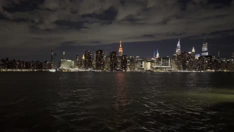 a captivating nighttime view of the new york city skyline, highlighted by the empire state building and its reflection on the east river, with dramatic clouds overhead adding to the scene's beauty