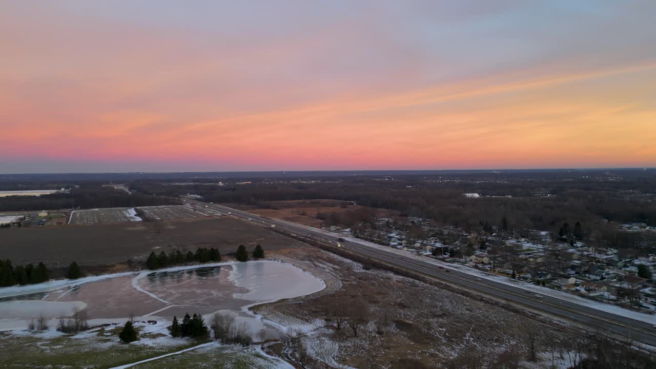 una autopista atraviesa un paisaje abierto al atardecer, el cielo brillando en tonos rosados y naranjas, con la escena evocando tranquilidad y movimiento