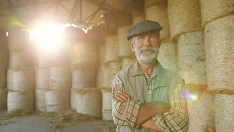 caucasian old gray-haired man farmer in hat standing in stable with hay stocks and looking at camera