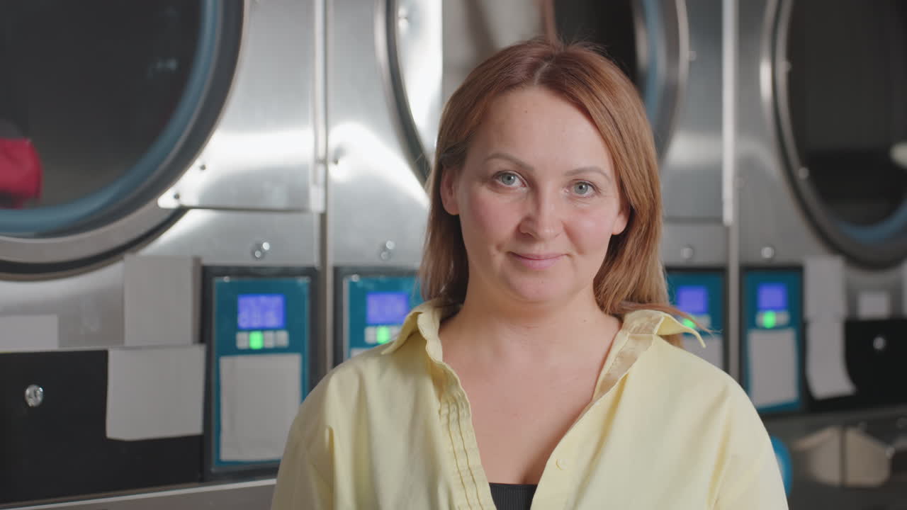 Portrait of happy mother standing before industrial laundry dryer and washer in modern laundromat, drums turning, digital panels glowing, clean facility vibe, service reliability, look to camera