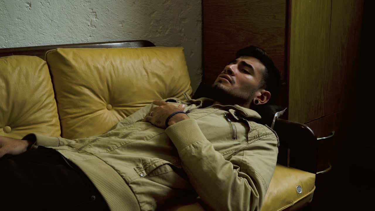 Distressed young man lying on a yellow leather couch, experiencing anxiety, agitation, and emotional pain in a dimly lit room