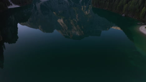 Snowy-limestone-peaks-mirroring-from-the-waters-of-lake-Pragser-Wildsee,-Dolomites,-Italy---Aerial-tilt-reveal