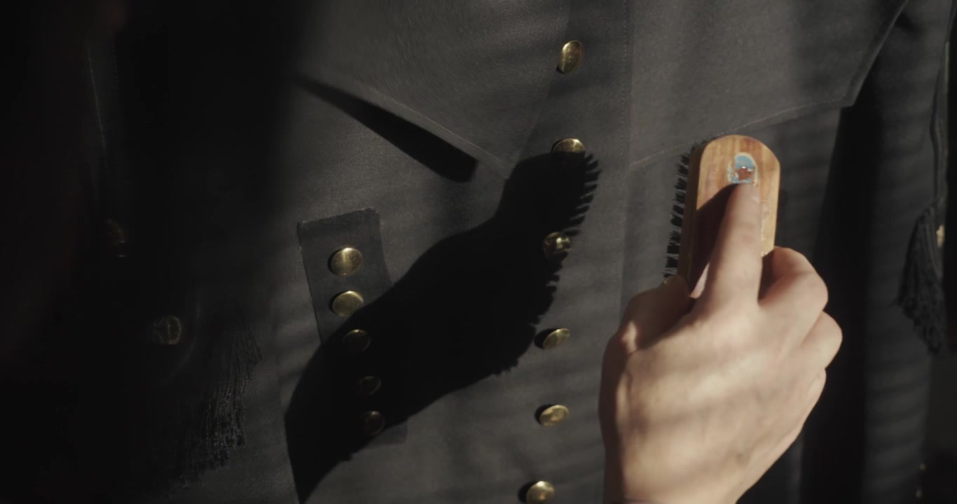 Wardrobe caretaker removes loose hairs and dust from the buttoned tunic of a traditional miner's ceremonial costume, demonstrating careful garment preparation and maintenance.