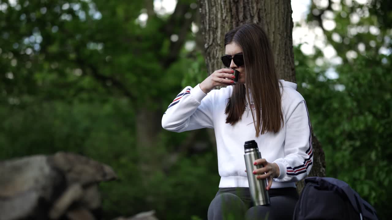 hermosa joven caucásica sentada en el bosque y bebiendo bebidas calientes de una botella de termo