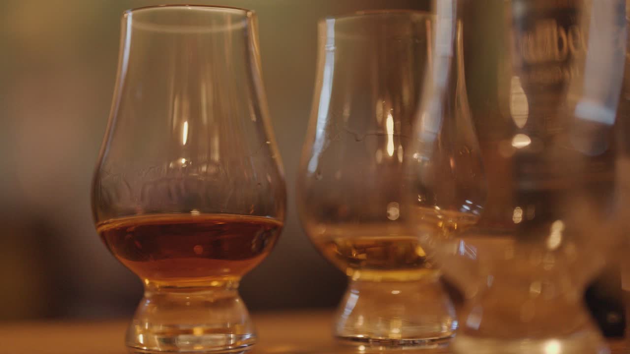 Three whiskey tasting glasses sit on a wooden bar, gradually shifting focus. Warm ambient lighting creates a cozy, inviting atmosphere with shallow depth of field
