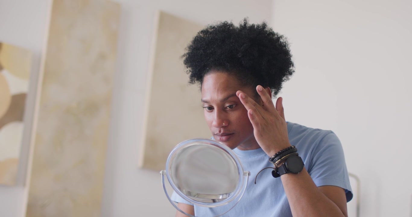 Uomo afroamericano che controlla l'occhio destro allo specchio della camera da letto, premendo sotto l'occhio e sistemandosi il lobo dell'orecchio.