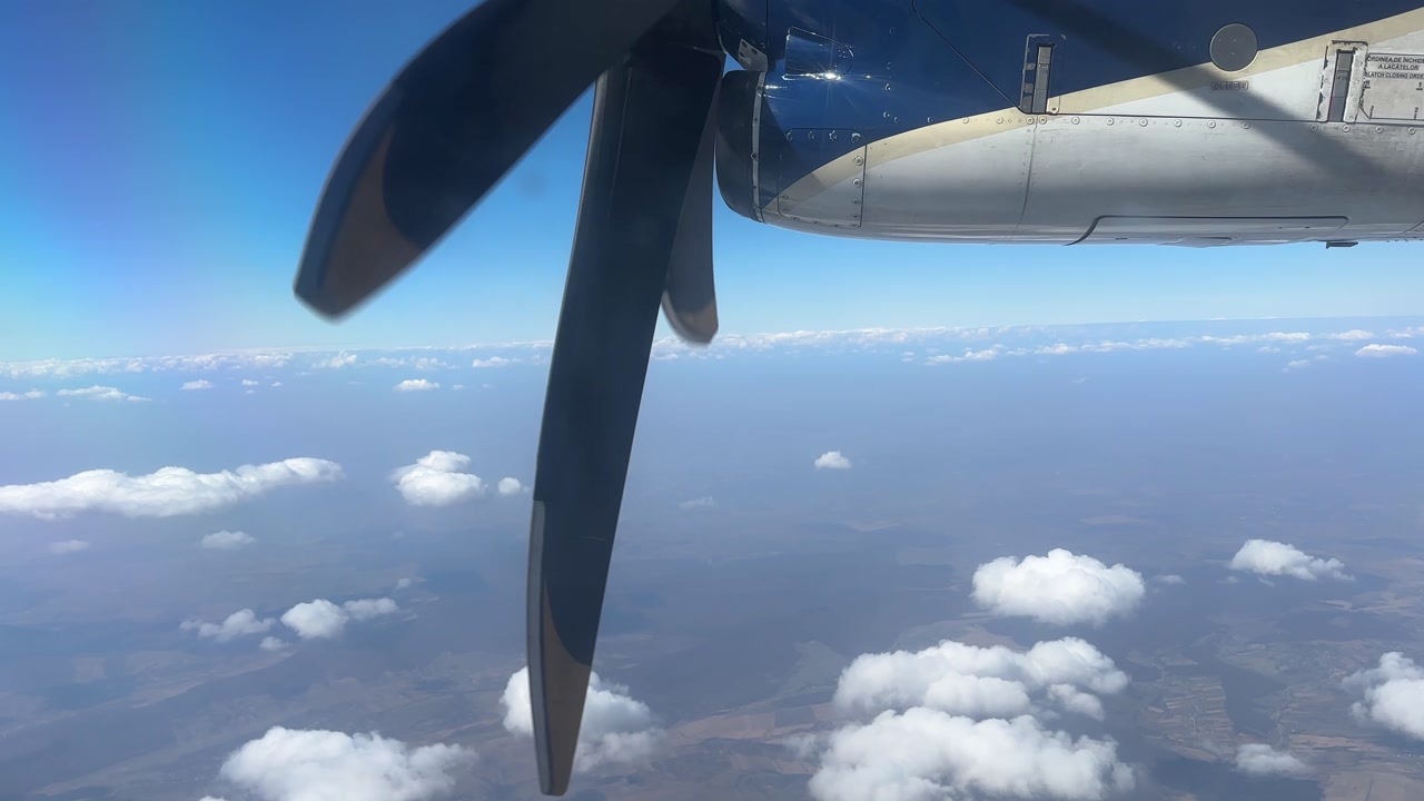 View from an ATR turboprop aircraft window showing the spinning propeller and engine above scattered clouds. Aerial perspective highlights flight experience and regional aviation travel