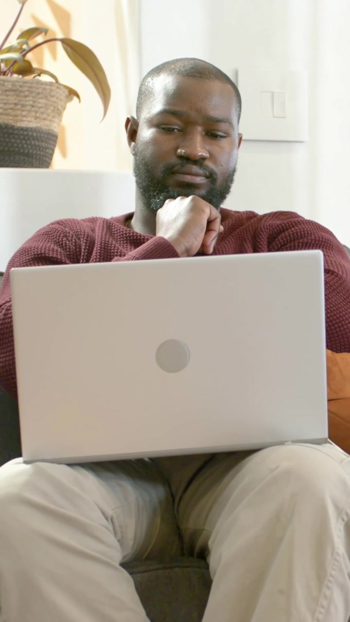 Vertical video: Sitting man resting chin, reviewing work on silver laptop at home, maroon sweater