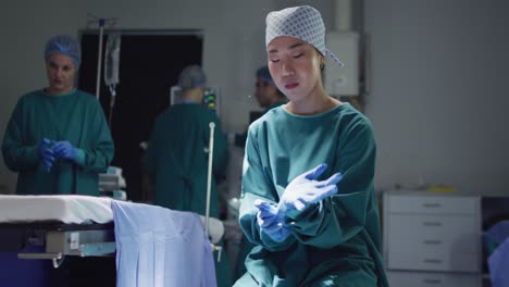 Video-of-asian-female-surgeon-putting-on-gloves-and-looking-to-camera-in-opreting-theatre