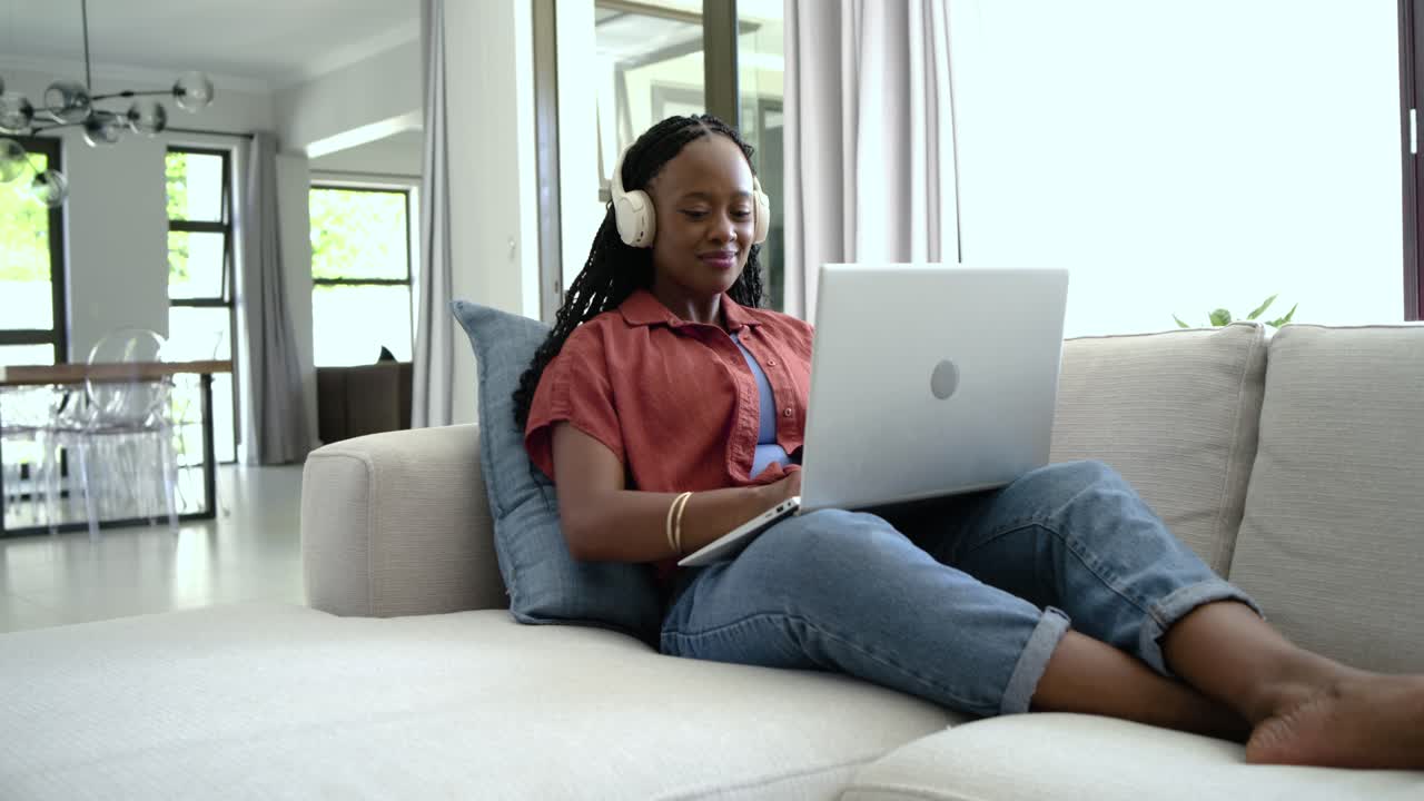 Hearing audio adult African woman sitting on sofa with headset typing on laptop smiling and working
