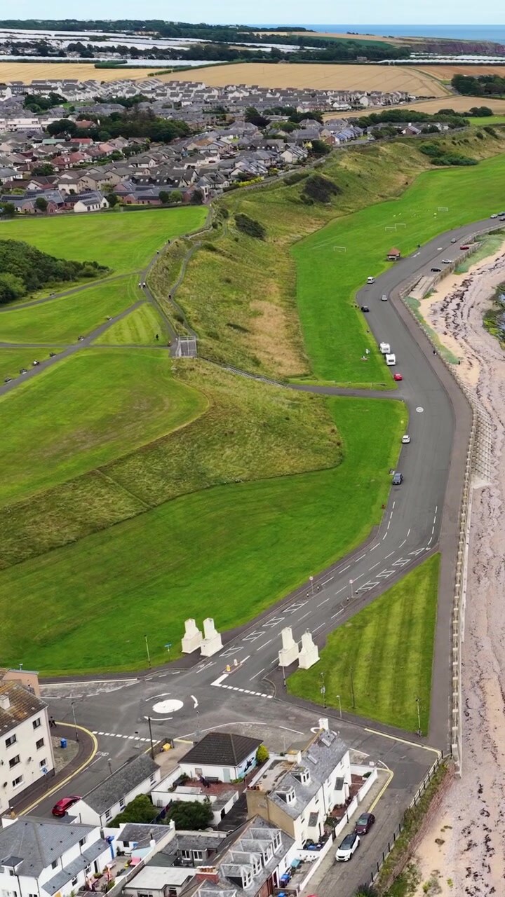 Smooth aerial drone shot over green fields, village houses, winding coastal road, and shoreline
