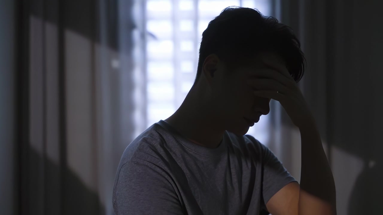 Distressed young husband man with hands on face looks worried and sad, showing stress, pain, headache, sadness, heartbreak, disappointmen, head bowed in deep thought anxiety, sorrow in dark bedroom