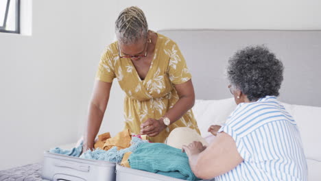 happy senior african american female friends packing suitcase on bed at holiday home, slow motion