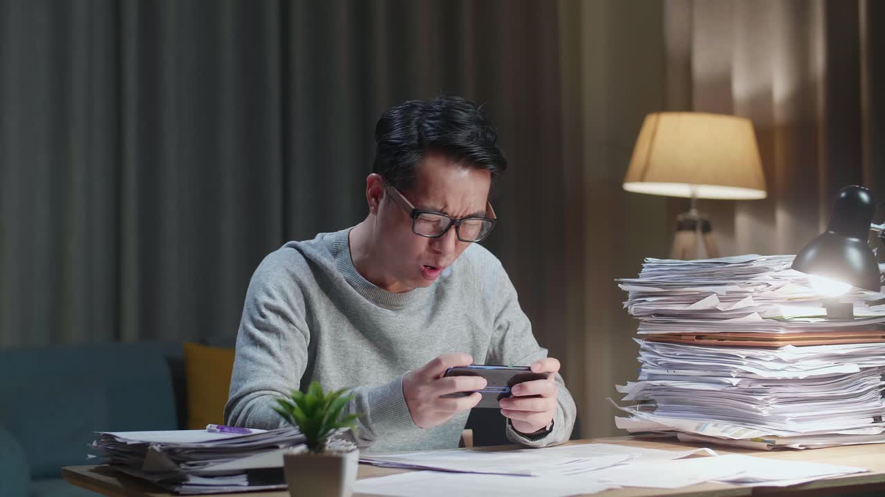 亚洲男子在家处理文件后，玩手机游戏输了，感到沮丧。