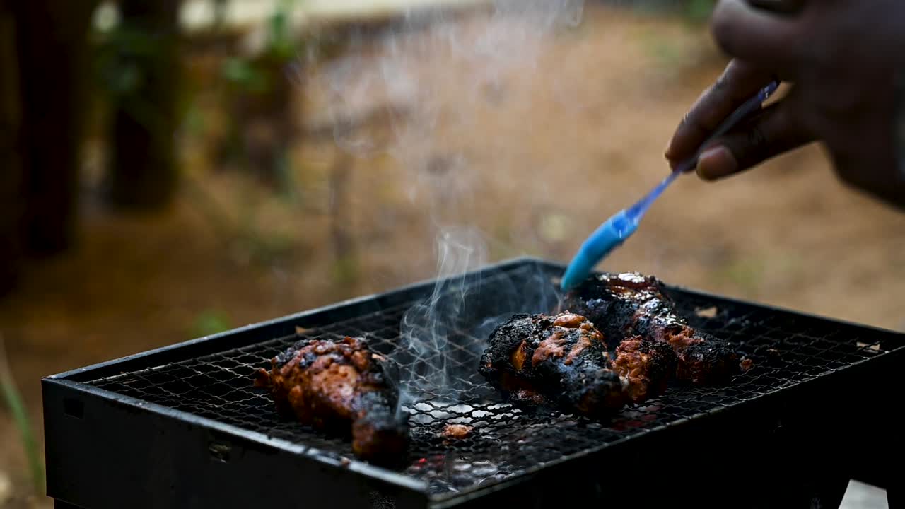 Primo piano al rallentatore di olio spennellato su cosce di pollo bruciate su una griglia per barbecue infuocata. Fiamme e fumo si alzano, catturando l'olio sfrigolante sulla carne carbonizzata e l'intensità della cottura all'aperto.