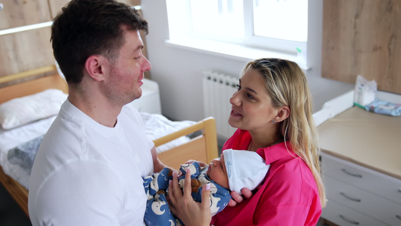 Caucasian parents stand in the hospital ward holding their baby. Happy couple talk smiling. High angle view.