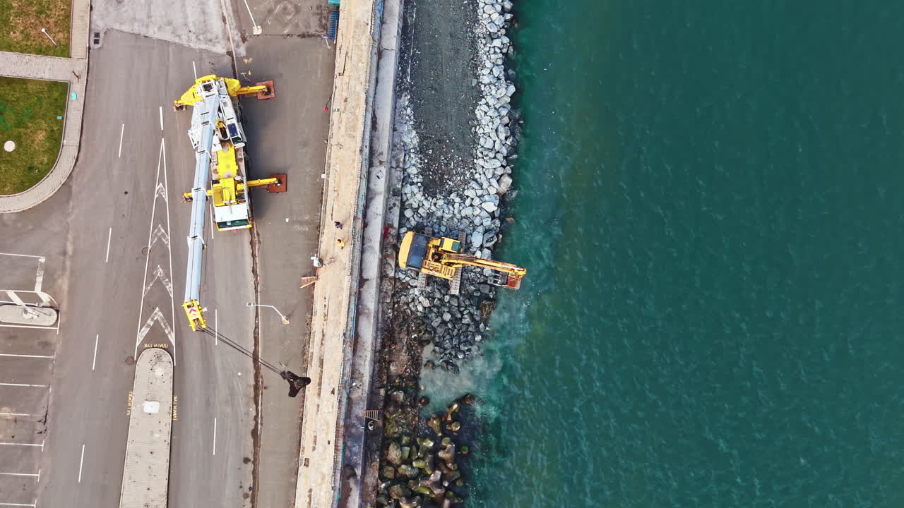 Lavori di costruzione si svolgono in un'area lungomare dove una gru e un escavatore spostano materiali lungo la costa durante le ore diurne.