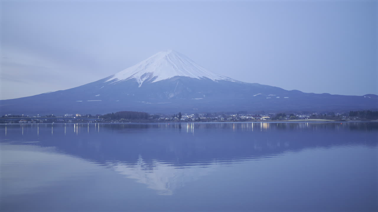 Il Monte Fuji si erge maestoso nel cielo crepuscolare del mattino, con il suo riflesso che luccica nelle acque calme del lago. I pittoreschi dintorni esaltano l'atmosfera tranquilla. Lago Kawaguchiko