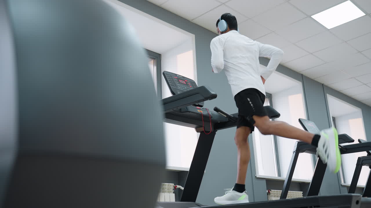 side view of man in white top and black shorts running on treadmill in bright fitness center with visible display screen, headphones on, showcasing energy, motion and determined