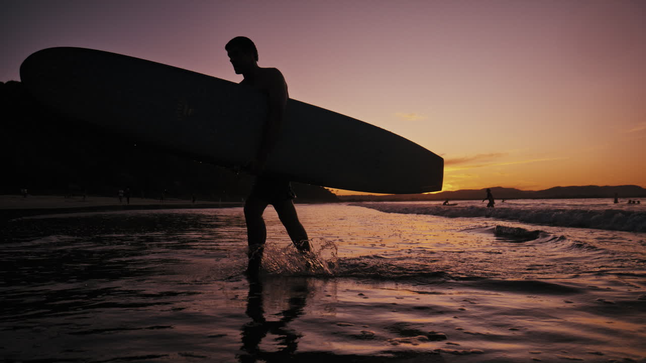 Surfer draagt board door ondiep water terwijl schemerkleuren zich over de lucht verspreiden, slowmotion silhouet