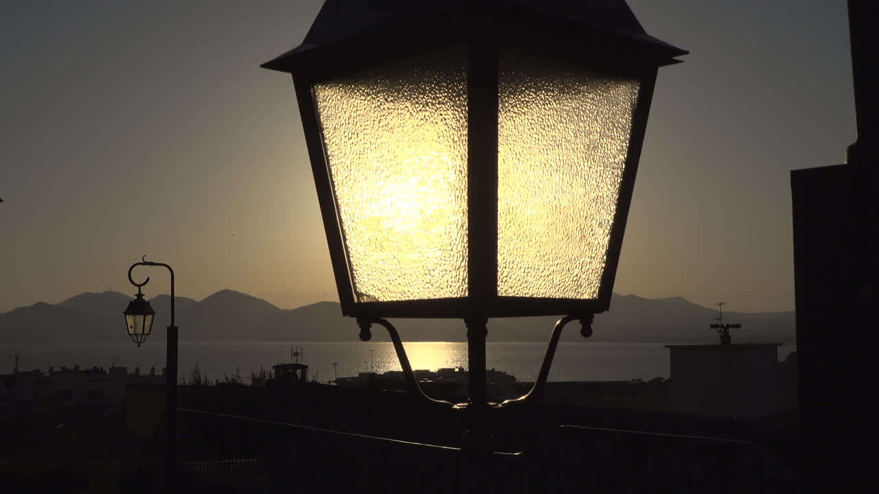 Close up of a lighted street lamp with the sea and the mountains on the background at sunset