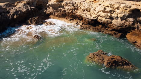 In-aerial-perspective,-the-rugged-coastline-of-Portugal-unfolds,-as-the-gentle-sea-waves-delicately-embrace-the-rocky-formations-under-the-radiant-sunshine-of-a-delightful-day