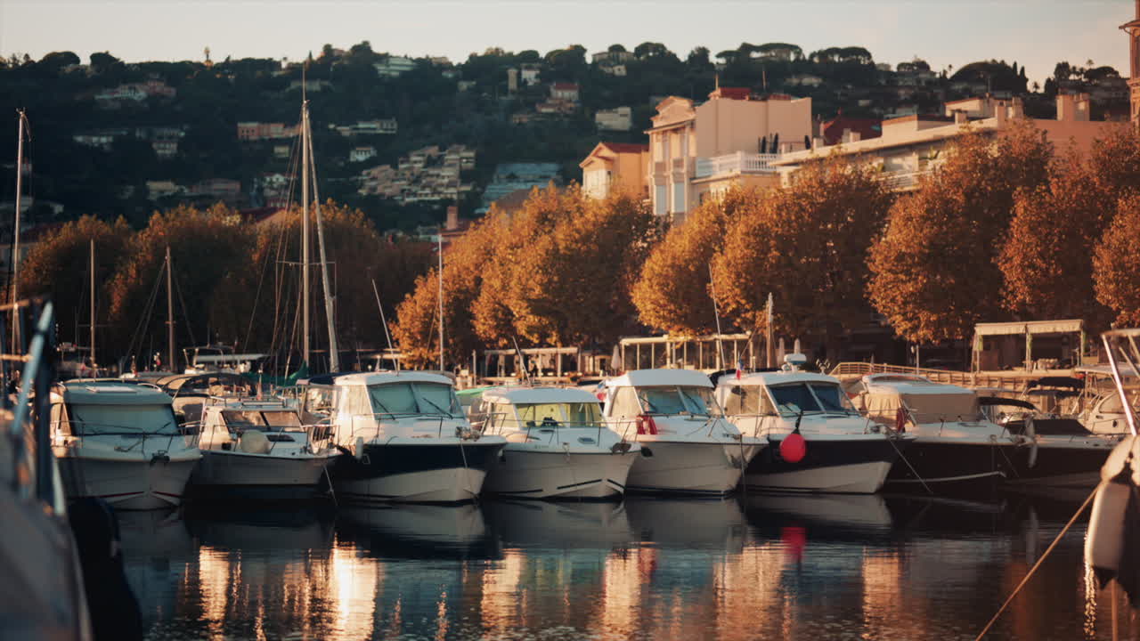 Row of boats docked near autumn trees and city buildings, reflecting golden evening light on calm water