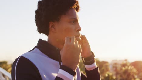 Close-up-of-african-american-man-putting-earphones-on-during-exercise-outdoors-by-seaside