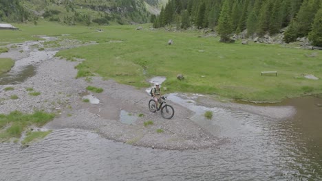 Aufsteigende-Luftaufnahme-Eines-Männlichen-Mountainbikers,-Der-Nach-Der-Ankunft-Am-See-In-Den-Bergen-In-Die-Drohnenkamera-Winkt