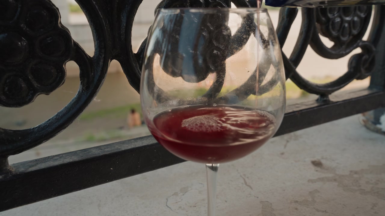 Liquido rosso viene versato in un bicchiere da vino trasparente su una superficie di cemento, vicino a una ringhiera in metallo nero decorata. Lo sfondo mostra vegetazione sfocata e figure distanti, suggerendo un ambiente esterno con luce naturale.