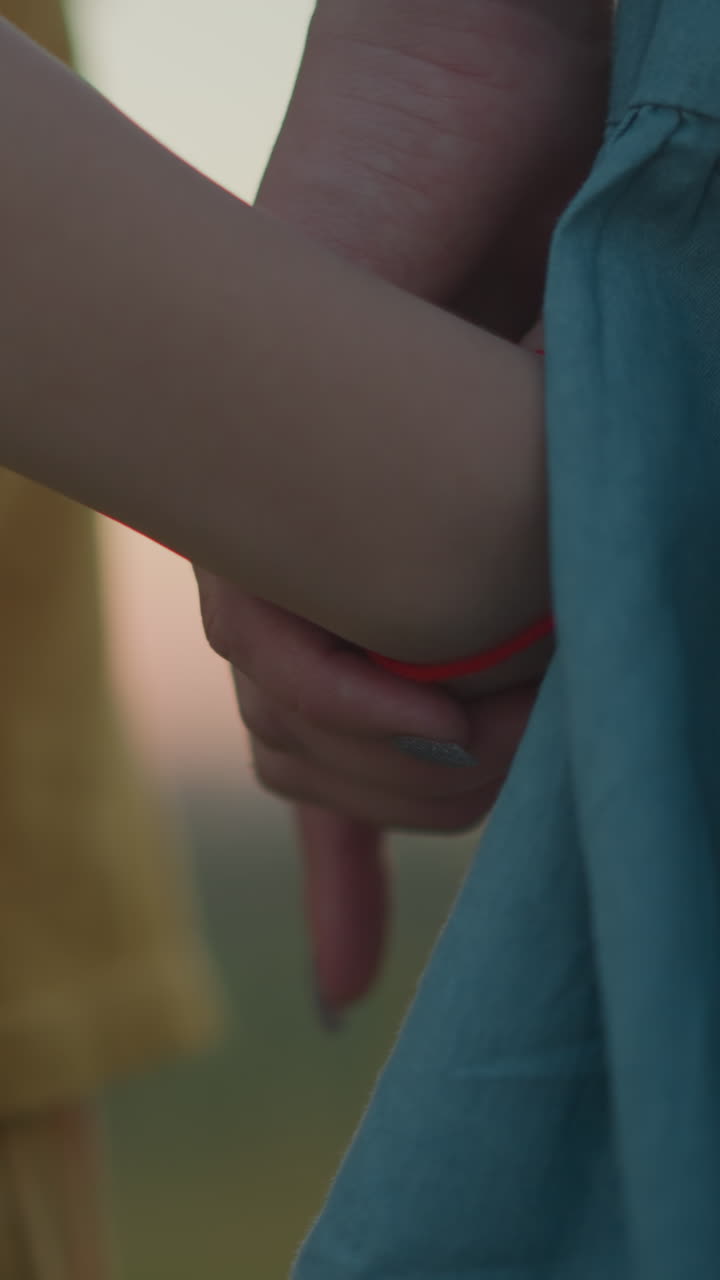 A touching close-up of a mother holding her son's hand, who is wearing yellow, as they stand together, facing away from the camera, watching the serene sunset over a tranquil lake