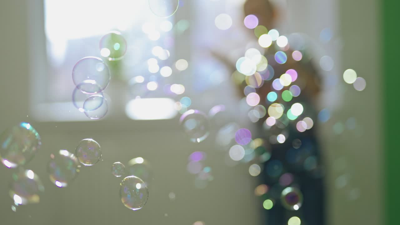 Multi-colored bubbles flying in the room on the blurred background with a boy. A lot of soap bubbles and a child who is playing with them at home. Close-up.
