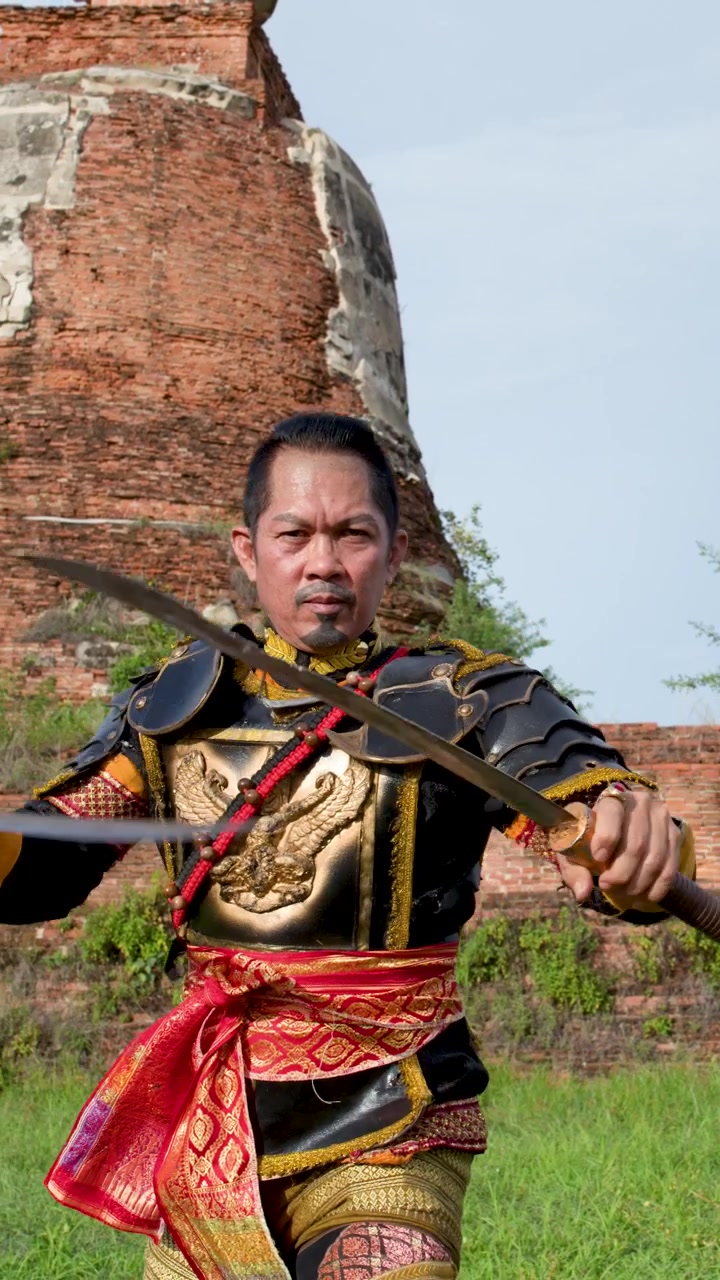 Seorang pria berzirah tradisional Thailand menampilkan seni bela diri dengan pedang di dekat reruntuhan bata kuno