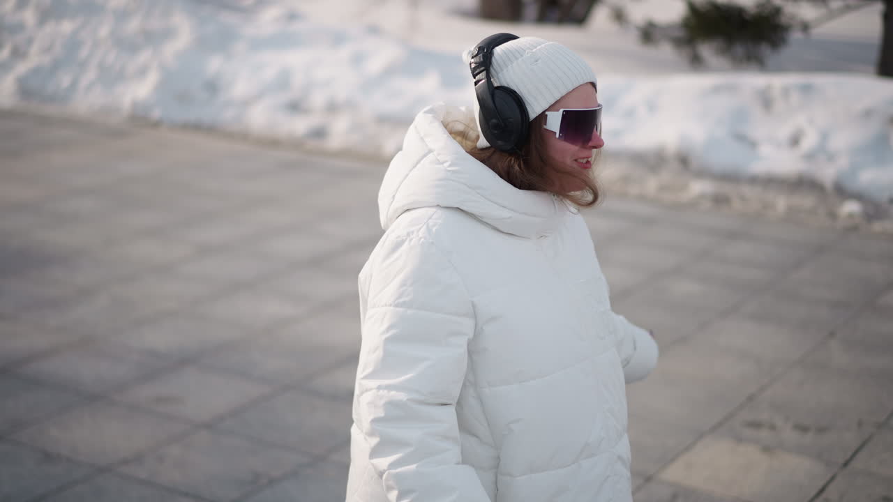 Adventurous school girl exercises with joyful energy on snowy winter plaza, moving arms in expressive dance like motions while wearing puffer jacket beanie and headset, smiling in excitement