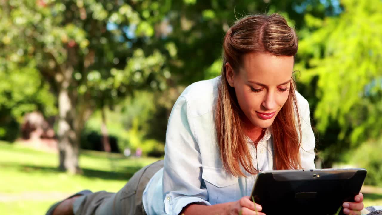 Pretty blonde using her tablet lying on a blanket in the park